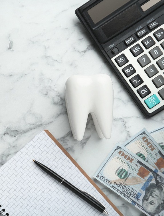 Model tooth next to $100 bills, calculator, and notepad with pen on marble surface